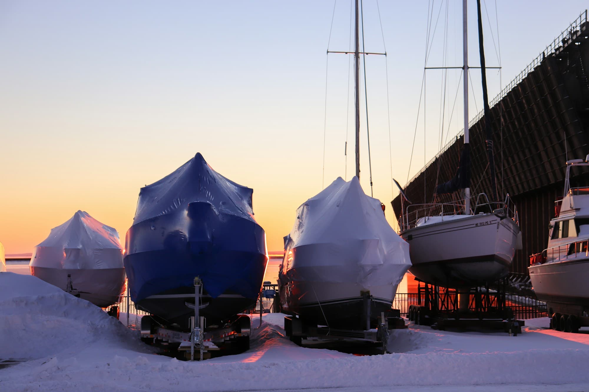 Boats shrink-wrapped in blue and white covers stored on trailers for winter at sunset